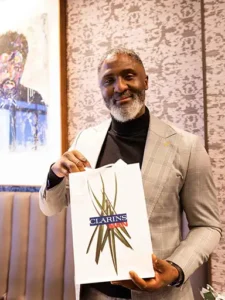 A man in a grey suit holding a Clarins-branded gift bag with a smile, standing in an elegant room with artwork.