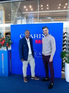 Clarins honours social fathers. Two men posing together in front of a blue Clarins backdrop, one in a navy blazer and beige pants, the other in a beige sweater and maroon pants, with floral arrangements on either side.