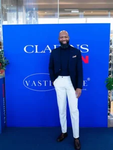 Clarins honours social fathers. A man in a dark suit and white pants standing confidently in front of a blue Clarins backdrop with floral decorations.