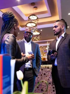 Clarins honours social fathers. Three individuals engaged in conversation at an event, with a woman in a leather jacket and two men in suits, standing near a decorated table.