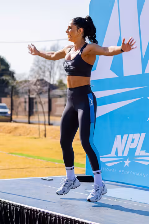 A fit woman in NPL branded activewear, mid-jump during an outdoor fitness class, with a blue and white NPL backdrop.