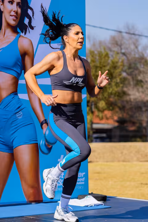 fitness instructor in NPL branded activewear vigorously exercising on an outdoor stage, with a large branded banner behind her.