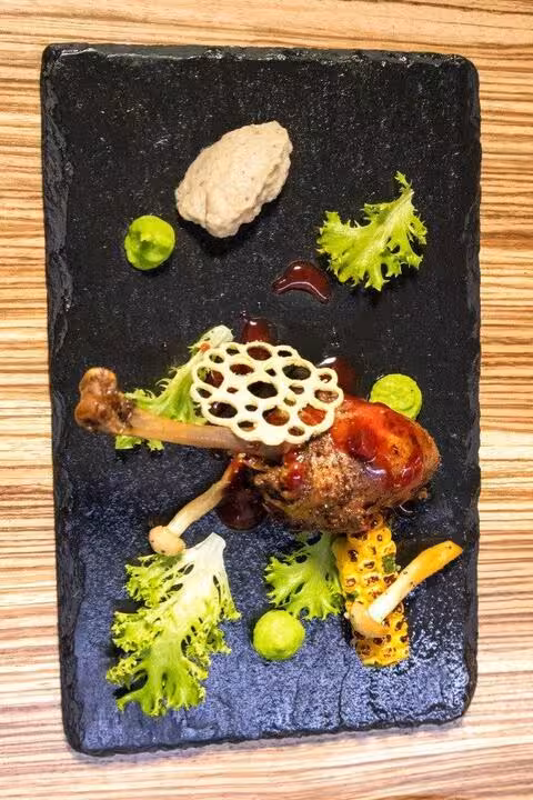 A confit duck leg, richly sauced and garnished with fresh greens, mushrooms, and grilled corn, elegantly presented on a dark slate serving board, demonstrating the artful plating and delicious main courses at Chef's Table Sandton Sun.