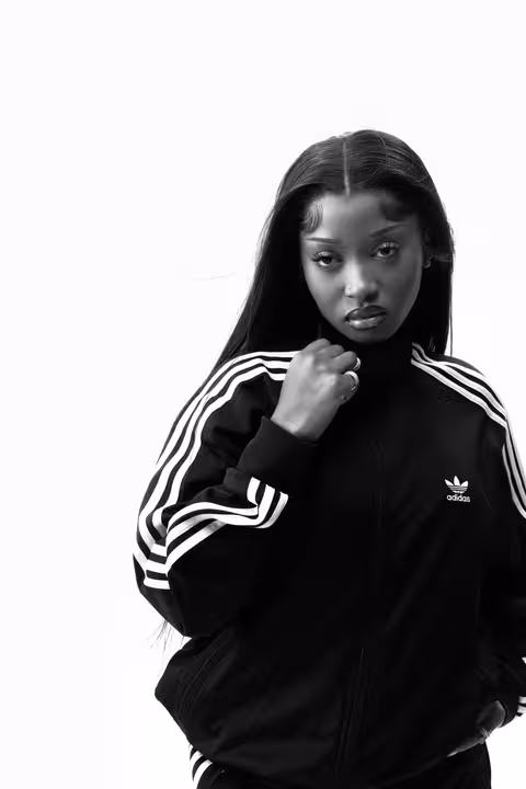 A black and white portrait of Uncle Waffles wearing a black adidas tracksuit jacket with white stripes, representing the Superstar campaign.