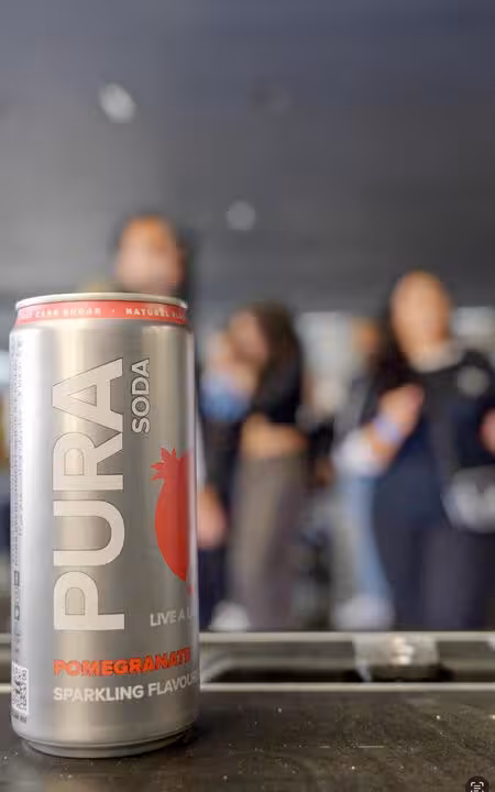 A can of PURA Pomegranate Sparkling Flavour soda is in sharp focus in the foreground, with a blurred background showing people at a social gathering, likely in Johannesburg. This highlights the beverage's presence within South Africa's wellness scene. The text "LIVE A LITTLE PURA" is visible on the can.