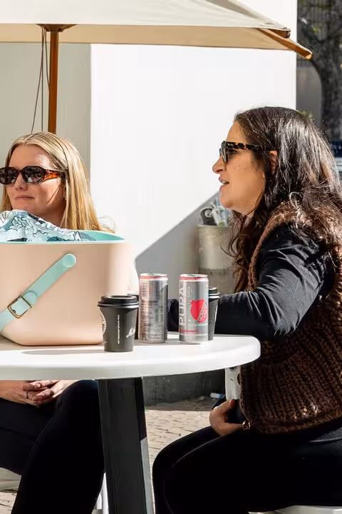 Two stylish women are seated at a white table outdoors in Johannesburg, under a light-coloured umbrella. There are cans of PURA soda and takeaway coffee cups on the table, suggesting a relaxed and wellness-oriented social gathering typical of South Africa's evolving scene.