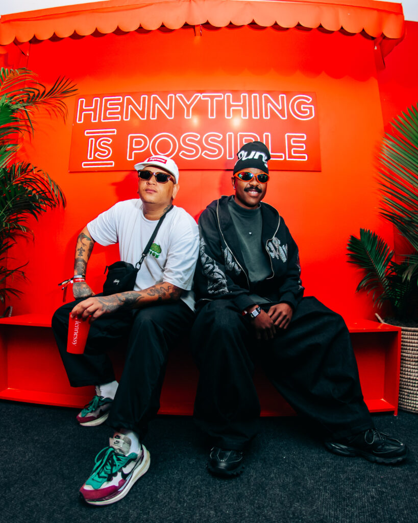 South African creatives African Ginger and Uncle Partytime sitting side-by-side on a red bench in front of the 'HENNYTHING IS POSSIBLE' neon sign.