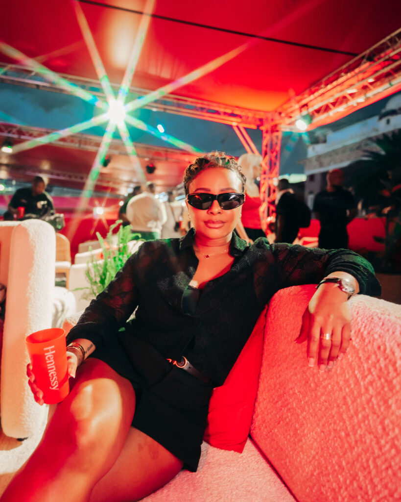 South African personality Boity Thulo relaxing on a plush pink sofa on the VIP deck, holding a red Hennessy cup.