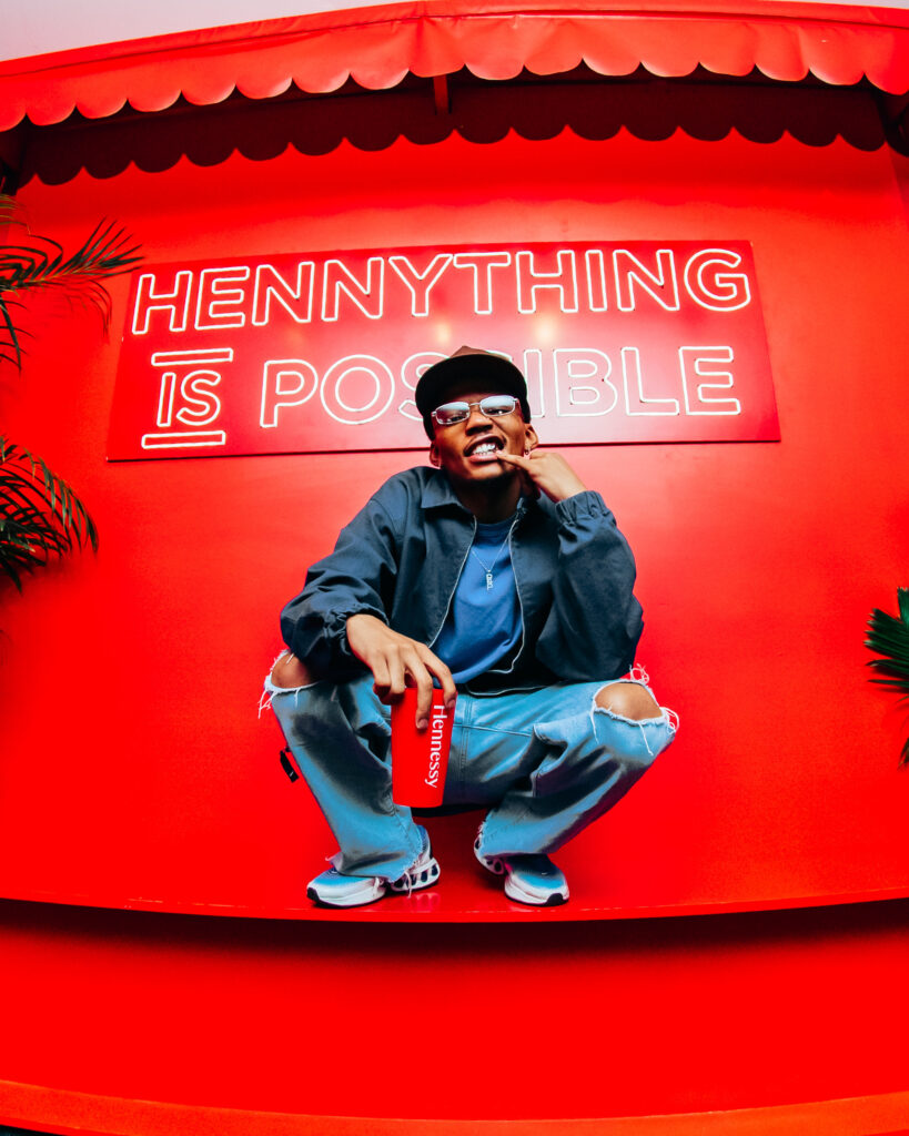 South African rapper Loatinover Pounds squats on a red platform holding a Hennessy can, posing in front of a neon sign that reads 'HENNYTHING IS POSSIBLE'.