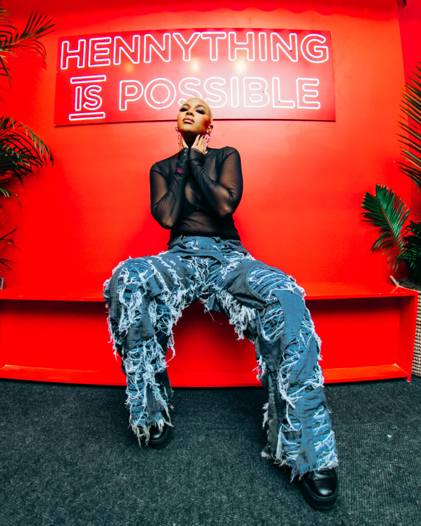 Media personality Luthando 'Loot Love' Shosha sitting on a red bench wearing distressed denim pants in front of the 'HENNYTHING IS POSSIBLE' neon sign.