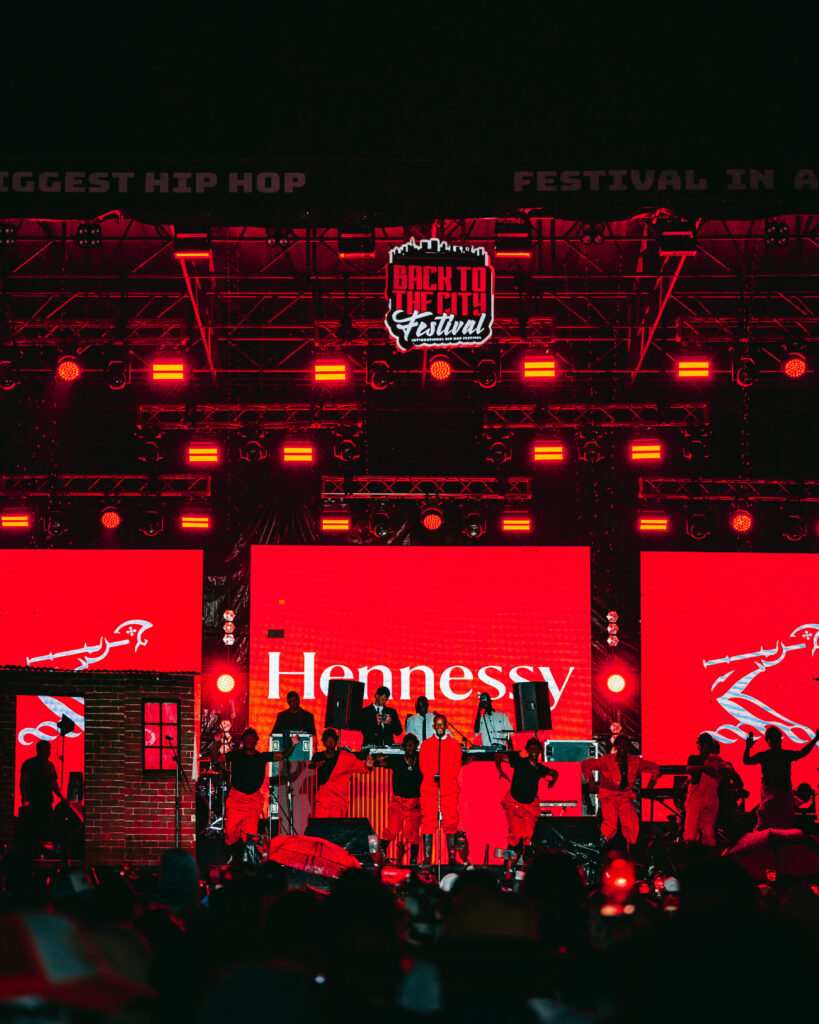 A wide shot of the main stage during a rainy performance at the Back to the City Festival, clearly showing the Hennessy and festival branding.