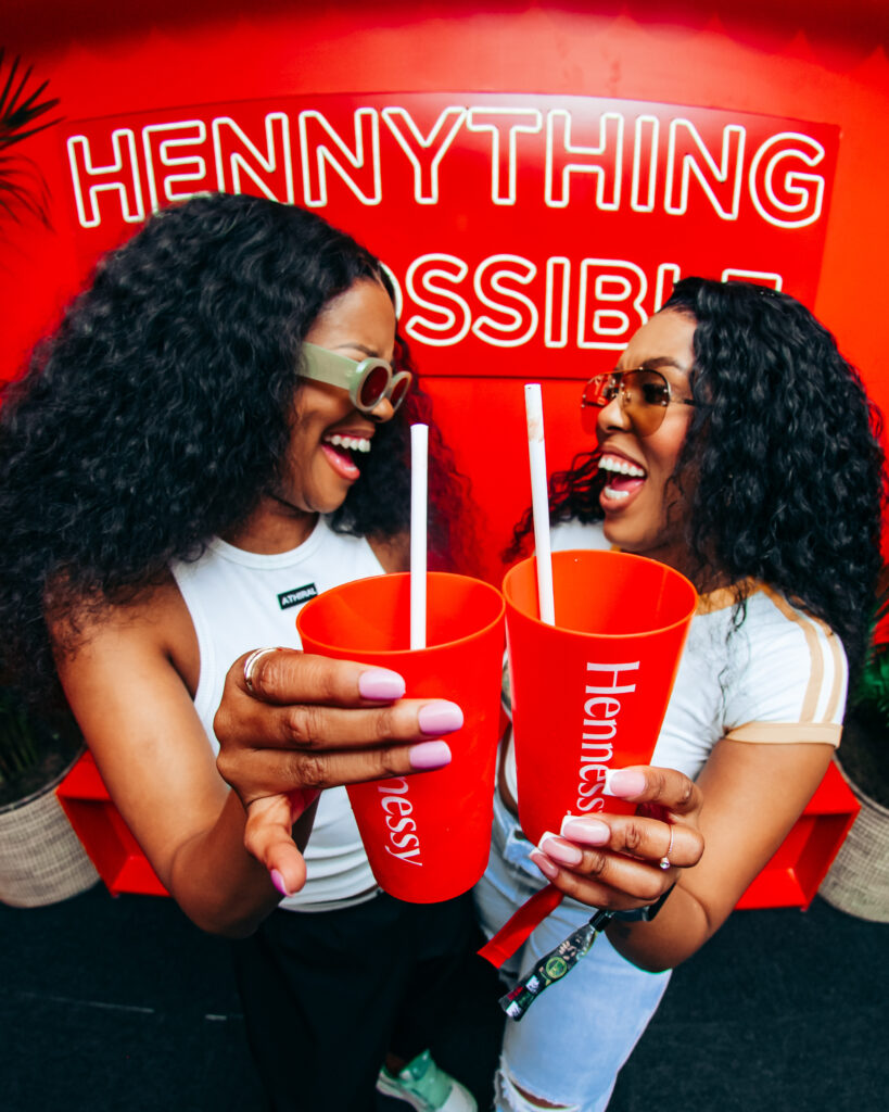 Influencers Neliswa Ngada and Naledi Malella clinking red Hennessy cups together and laughing in front of a red neon sign.