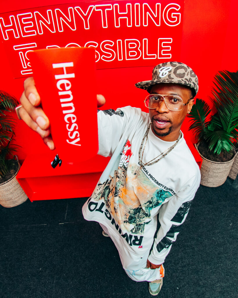 South African media personality Scoop Makhathini holding a red Hennessy cup, posing in stylish streetwear against a bright red 'HENNYTHING IS POSSIBLE' backdrop.