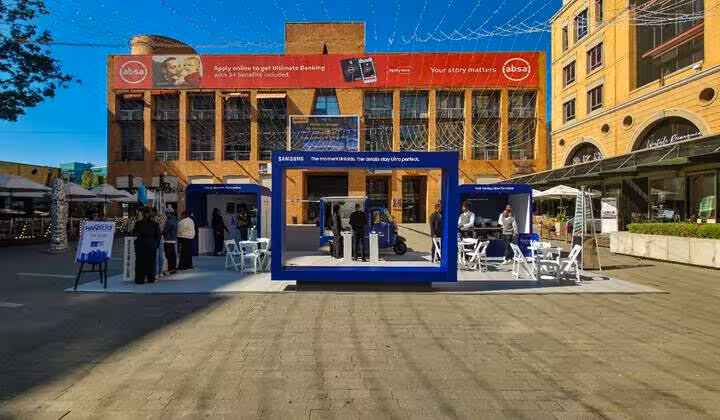 Absa bank branded building with a Samsung-branded blue pop-up experience booth in a busy outdoor Johannesburg square, possibly Nelson Mandela Square.