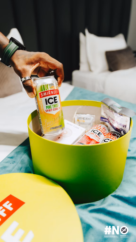 A hand places a Smirnoff ICE Pine Twist spirit cooler into a green welcome box filled with various curated items at the Sun City hotel room.