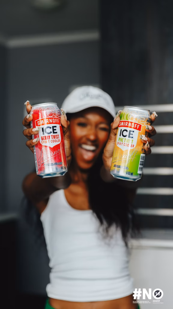 A young woman smiling while holding up two cans of Smirnoff ICE (Berry Twist and Pine Twist) directly towards the camera.